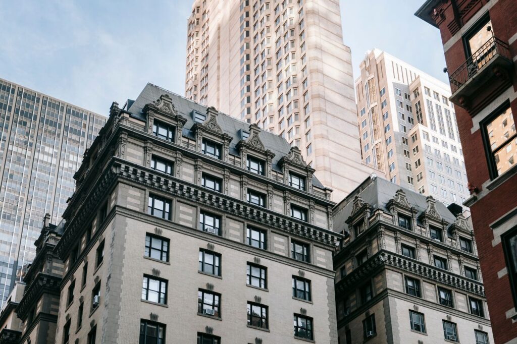 From below of residential building with white exterior and ornamented windows located near skyscraper against blue sky in city street